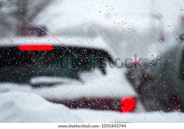 closeup of snowflakes and drizzle on windshield car\
driving through heavy winter city highway traffic with rear red\
lights of cars in front