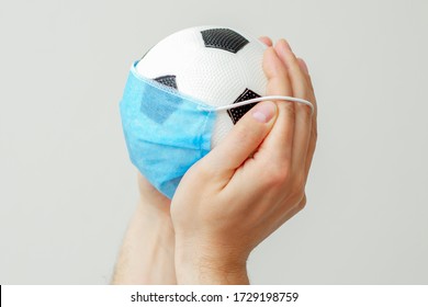 Closeup Of Soccer Ball With A Medical Mask In Man's Hands On A Light Background. Cancellation Of Sporting Events.