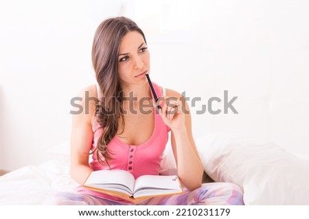 Close-up soft image pretty young woman sitting on her cozy bed and making notes in her diary in pink pajamas. Autumn season mood.