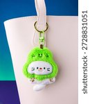 A close-up of a soft plush keychain featuring a cute and playful white kitten wearing a pear-shaped hat  on white bag
