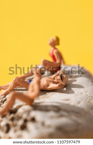 closeup of some different miniature people wearing swimsuit sunbathing on a rock, on a yellow background