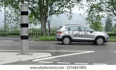 Close-up of a speed camera at a traffic light intersection with a silver-colored car driving past on a rainy, dreary day