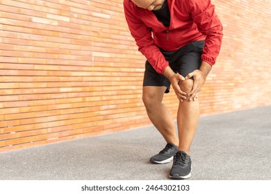 Closeup of sportsman wearing a red jacket injured his knee during exercise outdoors brick wall on city street. Athletic male in hoodie sweatshirt red suffering from joint pain in urban city park. - Powered by Shutterstock