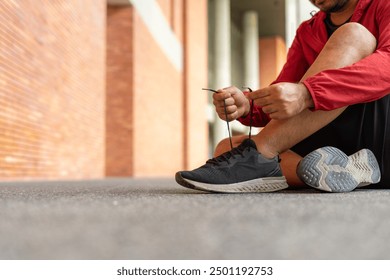 Closeup of sportsman wearing a red jacket tying sneakers outdoors brick wall on city street. Athletic male in hoodie sweatshirt red stopping lacing shoes in urban city park. - Powered by Shutterstock