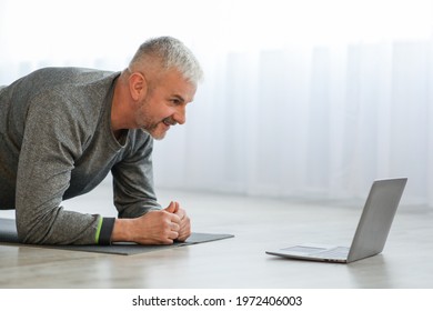 Closeup Of Sporty Senior Man Having Online Fitness Class While Staying Home During Coronavirus Pandemic, Planking On Fitness Mat And Looking At Laptop Scree, Side View, Copy Space