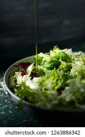 Closeup Of A Spurt Of Olive Oil Falling On A Mix Of Different Salad Leaves, Like Romaine Lettuce, Endive Or Arugula, On A Rustic Metal Dish