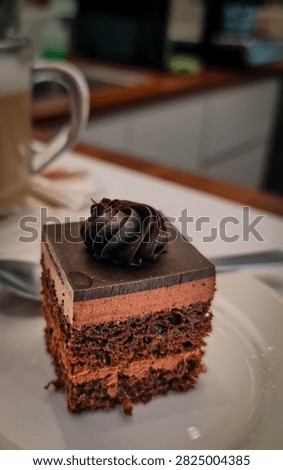 A close-up of a square slice of rich, multi-layered chocolate cake on a white plate. The cake features a smooth, glossy dark chocolate glaze on top, crowned with an elegant swirl of dark choc mousse or ganache. Below the glaze is a thick, creamy choc mousse layer, followed by moist, dense choc sponge cake layers interspersed with more cream filling. The cake has a deep brown color with visible texture, small air pockets, and embedded pieces, giving it a decadent, bakery-style appearance under soft lighting.