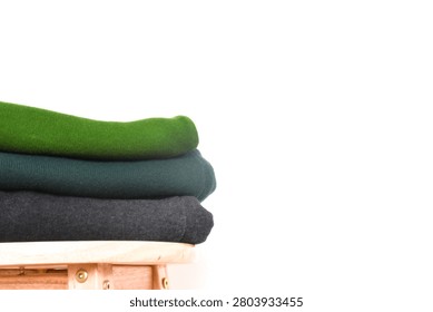 Close-up of a stack of folded green, dark green, and black clothes on a wooden stool against a minimalist white background