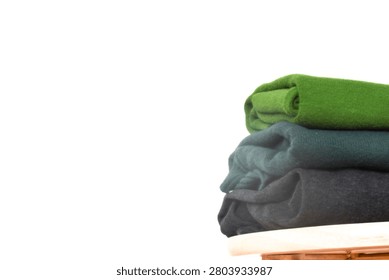 Close-up of a stack of folded green, dark green, and black clothes on a wooden stool on the right side against a minimalist white background