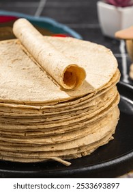 A close-up of a stack of fresh corn tortillas with one rolled tortilla on top, set on a black plate, showcasing traditional Mexican cuisine