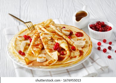 Close-up Of A Stack Of Freshly Cooked Sweet Crepes Topped With Cranberries, Banana Slices And Caramel Sauce On A White Plate On A Wooden Table, View From Above