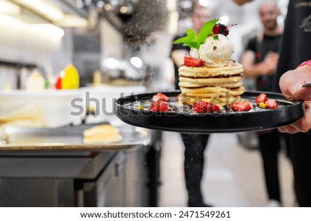 A close-up of a stack of pancakes with syrup, ice cream, and strawberries on a plate, garnished with a purple flower