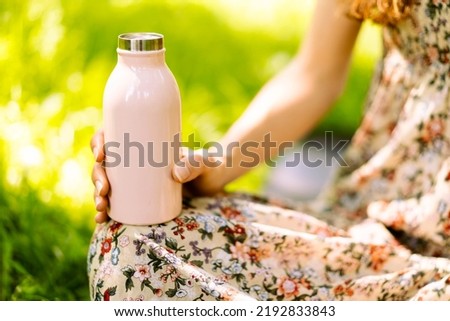 Close-up of steel eco thermo water bottle in female hand. Green background in the park. Copy space concept. Space for text. Plastic free. Zero waste concept. Selective focus. 