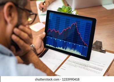 Closeup of a stock market broker working with graphs on digital tablet at office. Rear view of stock agent reading bad report and graph. Back view of multiethnic businessman analyzing fall sales. - Powered by Shutterstock - Get 15% off with code: PIKWIZARD15