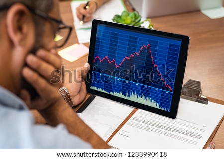 Closeup of a stock market broker working with graphs on digital tablet at office. Rear view of stock agent reading bad report and graph. Back view of multiethnic businessman analyzing fall sales.
