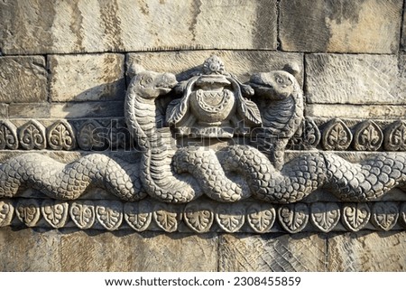 Closeup of stone carving ornaments on a Nepali temple, representing two intertwined snakes surrounded by lotus petals and holding the vase of jewels, one of the 8 auspicious symbols in Buddhism