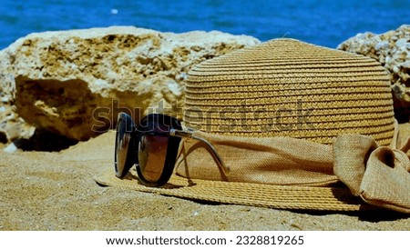 Close-up of straw hat, sunglasses on a sandy beach on a tropical coast on a warm sunny day. In the background are rocks and a calm Mediterranean sea with blue water. The concept of summer holidays
