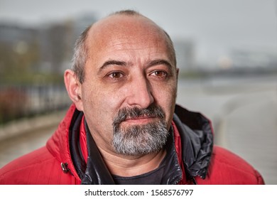 Close-up Street Portrait Of An Elderly Balding Man With A Gray Beard In Warm Clothes. The Face Of A Middle-aged Male Older Than 50 Years Outdoors. Fifty Year Old Businessman Outside In Cold Weather.