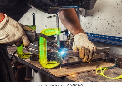 Close-up A Strong Man Welder In A Black T-shirt, Welds A Metal Welding Machine In The Garage In The Welding Mask, Blue Sparks Fly To The Sides, Protective Glasses And Tools Are On The Table