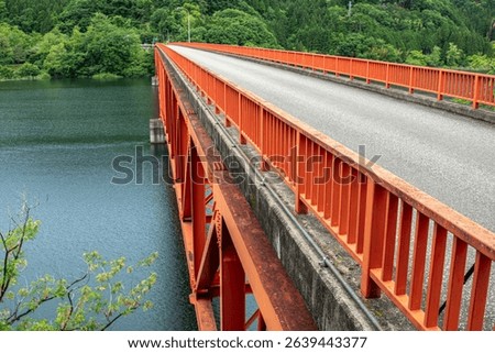 Close-up structural view of the red Kusaki Bridge spanning Kusaki Lake (Kusaki Dam) in Midori City, Gunma, Japan, showcasing its steel architecture against a backdrop of lush forest and calm water.

