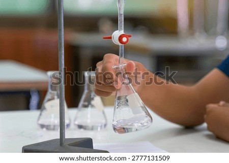  Close-up of a student's hand performing a titration experiment with a burette and Erlenmeyer flask in a chemistry lab. Concept of scientific research, education, and laboratory testing.                              