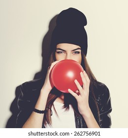 Closeup studio portrait of beautiful trendy hipster teenage girl blowing a red balloon wearing black leather jacket and black beanie hat. Square format, instagram look filter.