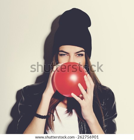 Closeup studio portrait of beautiful trendy hipster teenage girl blowing a red balloon wearing black leather jacket and black beanie hat. Square format, instagram look filter.