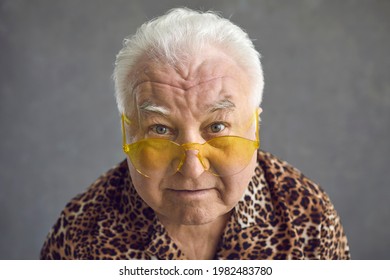 Closeup Studio Portrait Of Stylish Senior Man In Yellow Sun Glasses And Leopard Patterned Outfit. Old White Haired Guy With Chubby Face In Trendy Sunglasses Looking At Camera. Fashion And Style