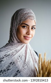 Close-up Studio Portrait Of A Tall, Slim And Elegant Middle Eastern Muslim Woman In A Black Dress And Hijab Head Scarf Against A Brown Background. She Is Holding A Bushel Of Wheat.