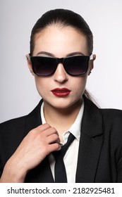 Close-up Studio Portrait Of Woman With Sunglasses, Red Lipstick And Suit Looking To Camera. Model With Conservative Hairstyle And Wearing Black Classic Suit With Tie And White Shirt