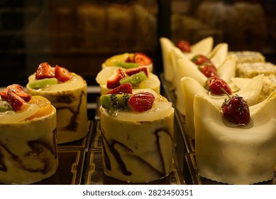 A close-up studio shot of assorted mini fruit cakes and white chocolate pastries displayed inside a bakery showcase cabinet.