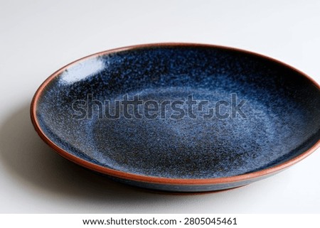 A close-up studio shot of a deep blue ceramic plate with a speckled texture and a rustic orange-brown rim, set against a plain white background.