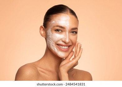 Close-up studio shot of happy European woman applying face mask on half of her face, posing against beige background, smiling at camera - Powered by Shutterstock