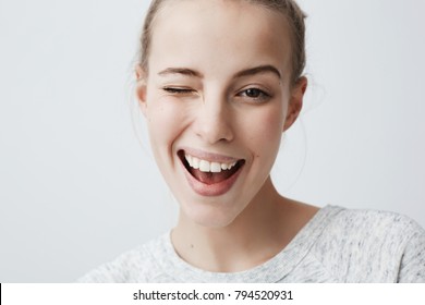 Close-up studio shot of positive coquettish young European woman with dark eyes, smiling happily, blinking at camera in a playful manner, flirting with you. Human facial expressions and emotions