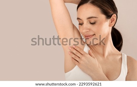Closeup studio shot of young smiling beautiful woman wearing white top touching her fresh hairless armpit, isolated on beige studio background, panorama with copy space. Hyperhidrosis treatment