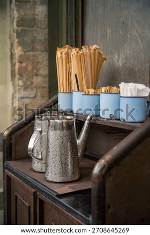 A close-up, stylized shot of a rustic coffee condiment station featuring vintage metal kettles covered in condensation and blue enamel mugs holding napkins, straws, and stirrers.
