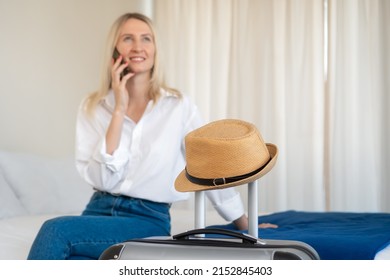 Close-up of a suitcase with a hat on the background of a smiling young woman in a white shirt, blue jeans sitting on a bed and talking on the phone. Concept of tourism, travel, happy life