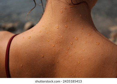 Close-up of a sun-kissed girl's back, adorned with glistening droplets, partially revealing strands of hair, with a backdrop featuring the sea. Emphasis on the droplets on the skin.