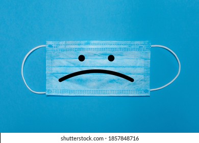 Close-up of a surgical mask on a blue background with a sad face. Concept of sadness caused by Covid-19, new normal and Blue Monday.