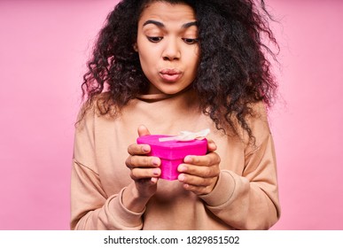 A Close-up Of A Surprised Smiling Girl With Dark Skin And Curly Lush Hair, Wearing A Beige Jacket, Holding A Hot Pink Heart-shaped Gift Box With A White Bow And Waiting To Receive A Long-awaited Gift.