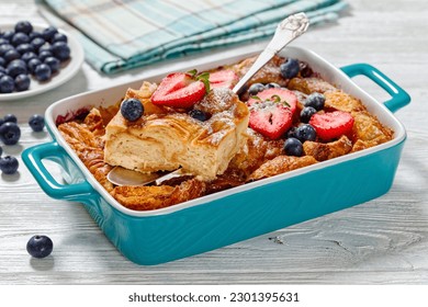 close-up of Sweet Croissant Breakfast Casserole with Berries in blue baking dish on white wood table with cake shovel, horizontal view from above