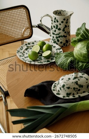 Close-up of table with green patterned ceramic jug and plate, fresh limes, cabbage, and leek next to rattan chair.