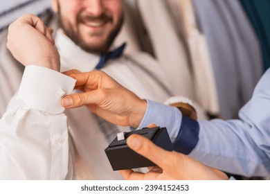 Close-up of a tailor fitting cufflinks on a client's shirt sleeve, both smiling at a wedding store - Powered by Shutterstock