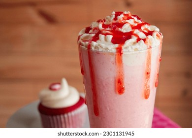Close-up tall glass filled with a fresh strawberry milkshake, topped cream with additional strawberries jam for the perfect refreshing dairy dessert. Added red velvet cupcakes behind