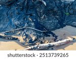 Close-up of the tar streaked rocks at Tar Pits Park in Carpinteria, California, showing a mix of sand, natural tar deposits, and driftwood on the shoreline