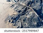 Close-up of the tar streaked rocks at Tar Pits Park in Carpinteria, California, showing a mix of sand, natural tar deposits, and driftwood on the shoreline