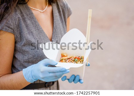 Closeup tasty food wok in box delivery. Girl holds udon noodles with tempuru, shrimps, soy sauce in hands in medical gloves. Japanese spicy food in disposable eco boxes. Wok box udon advertise
