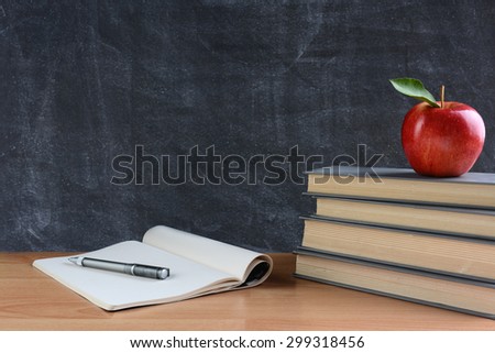 Closeup of a teachers desk with books, paper and pen and a red apple in front of a chalkboard. Horizontal format with copy space. 