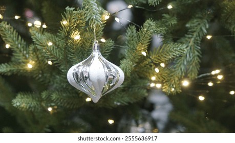 A close-up of a teardrop silver and white striped bauble hanging on a Christmas tree, illuminated by beautiful bokeh fairy lights in the background, creating a festive and elegant holiday scene. - Powered by Shutterstock