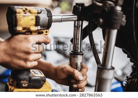 Close-up of a technician's greasy hands using an electric impact driver tool to loosen bolts on a motorcycle front suspension system during overhaul and repair in a garage workshop.